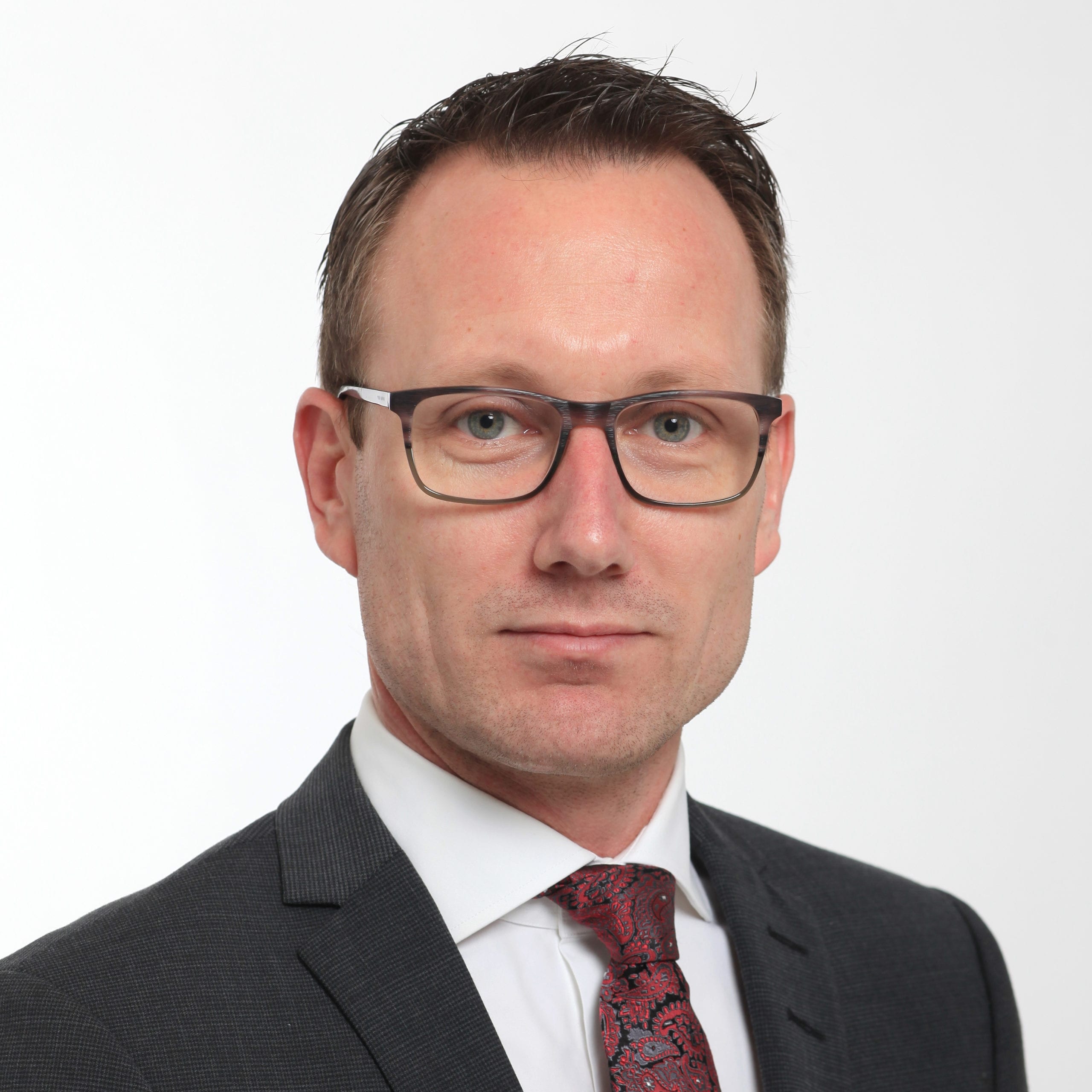 Headshot of a man wearing glasses and a dark grey suit with a white shirt and red tie.
