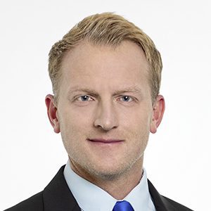 Headshot of Jacob Nico with short blonde hair and wearing a black suit with a blue tie.